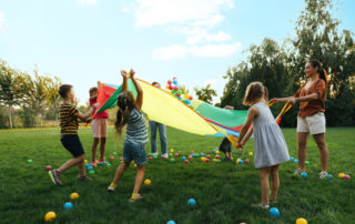 Group of children and teacher playing with rainbow playground parachute on green grass. Summer camp activity