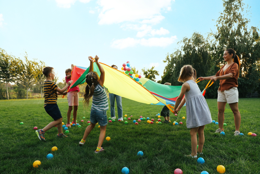 Group of children and teacher playing with rainbow playground parachute on green grass. Summer camp activity